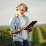 Agricultor idoso faz anotações em um caderno enquanto observa uma plantação verde.