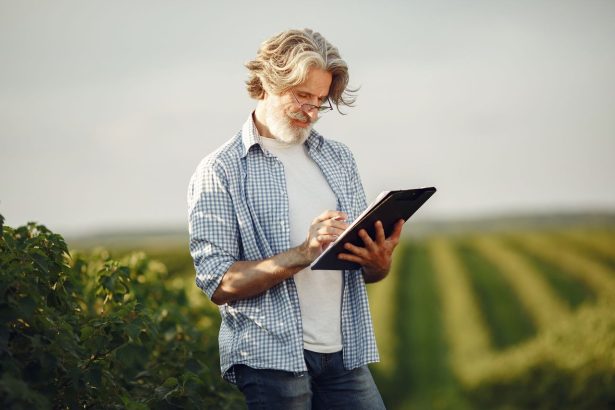 Agricultor idoso faz anotações em um caderno enquanto observa uma plantação verde.