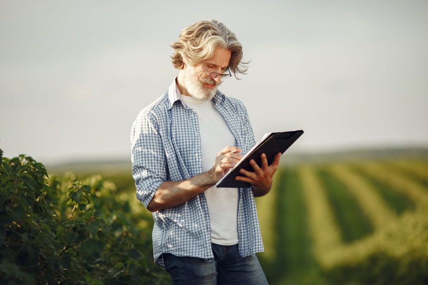 Agricultor idoso faz anotações em um caderno enquanto observa uma plantação verde.