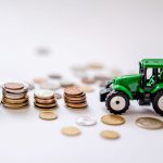 Miniatura de um trator verde ao lado de pilhas de moedas em uma composição sobre agricultura e finanças.