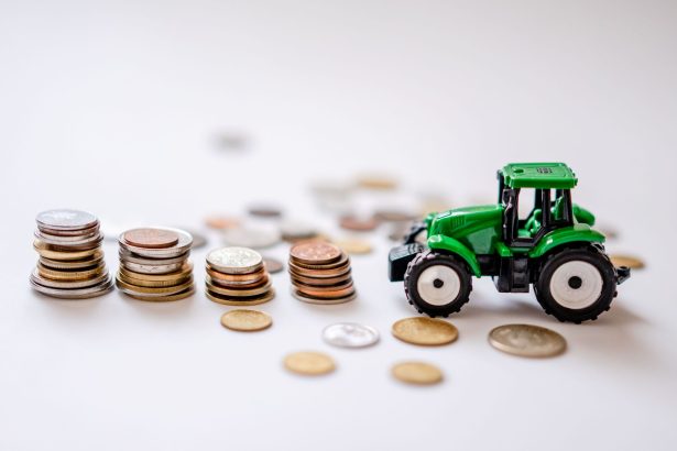 Miniatura de um trator verde ao lado de pilhas de moedas em uma composição sobre agricultura e finanças.