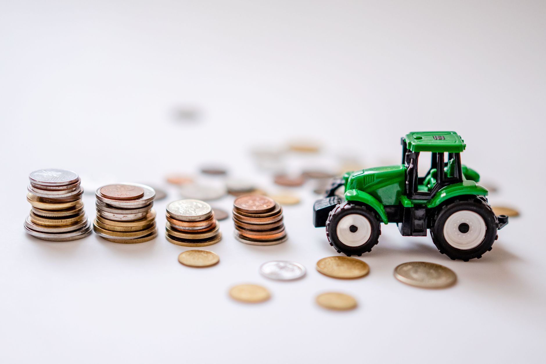 Miniatura de um trator verde ao lado de pilhas de moedas em uma composição sobre agricultura e finanças.